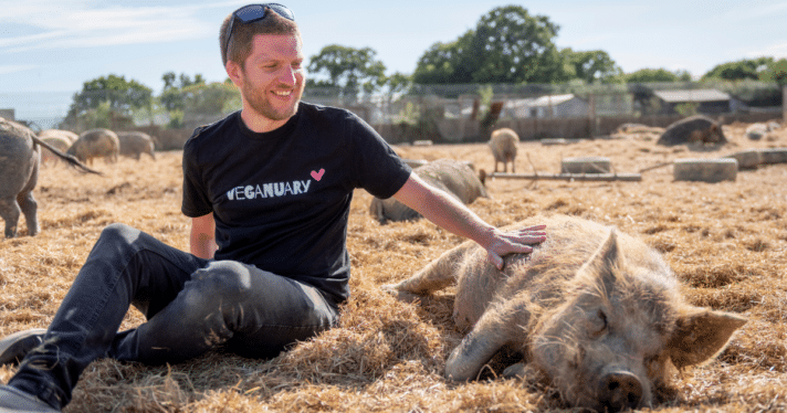 A man in a Veganuary shirt sitting with and petting a pig
