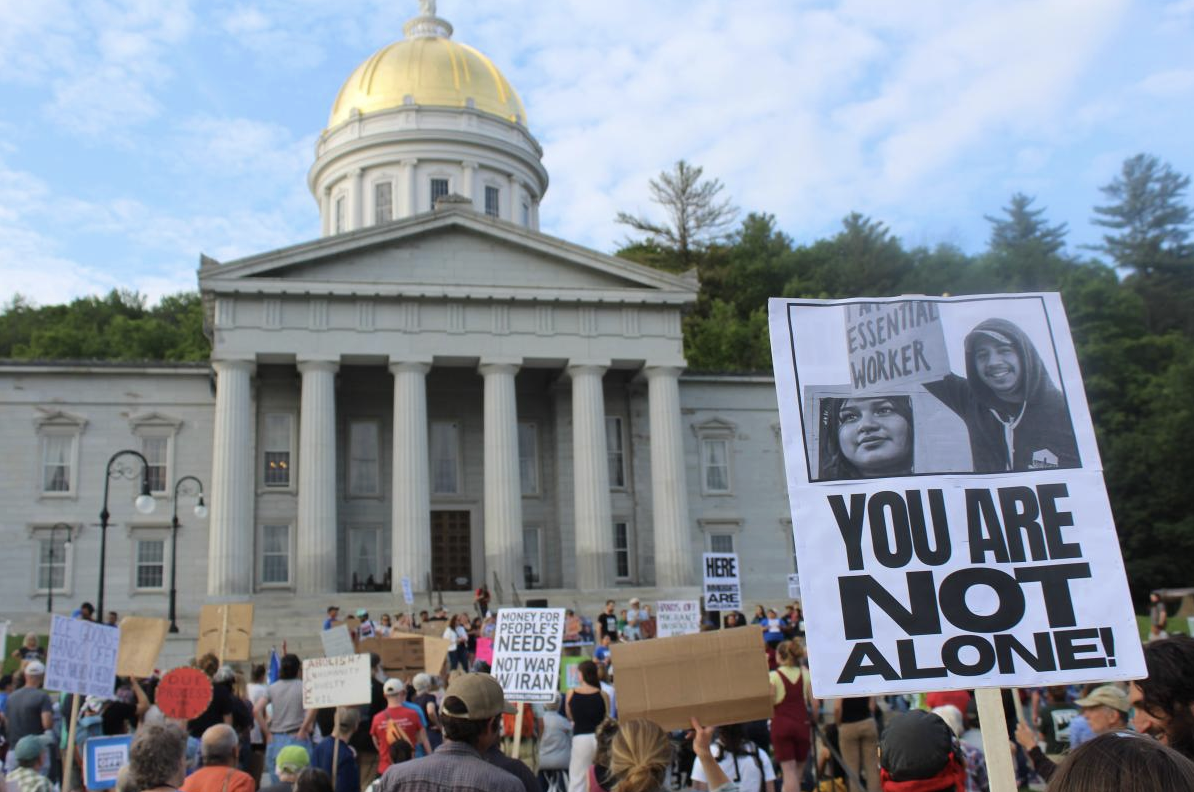 A protest outside the Vermont State House. A sign reading YOU ARE NOT ALONE! is facing the camera.