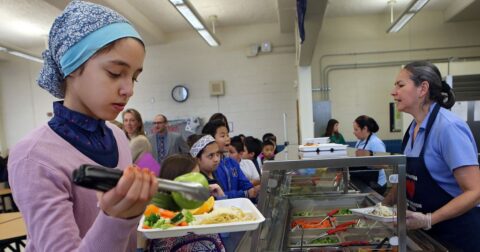 A child putting a green apple on their plate in line at a school cafeteria. Cafeterias are one place groups are exploring the power of nudges