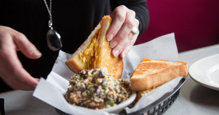 A person's hand lifting a sandwich next to a small bowl of caramelized onions