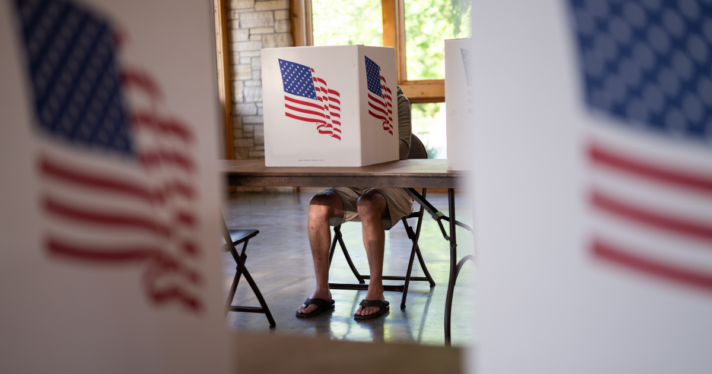 A person sitting behind a voting booth with an American flag on it
